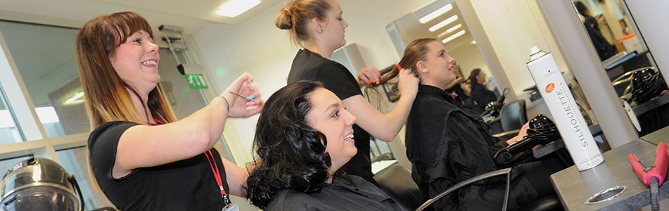 two women sat in chair having their hair styled by students